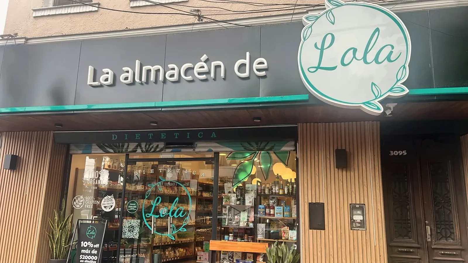 Fachada de La Almacén de Lola: cartel con el logo, vidriera iluminada con góndolas de madera y plantas en la entrada